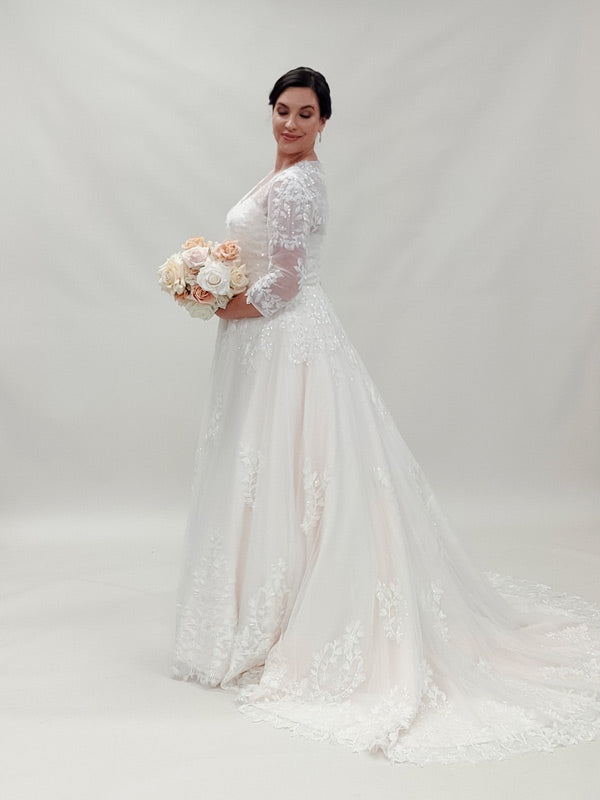 "Radiant plus-size bride in an ornate lace A-line lace wedding dress with a graceful train, holding a pastel rose bouquet"