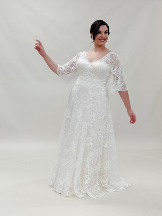 A radiant bride standing confidently in a boho wedding dress with a V-neckline and removable lace sleeves,  The Blair bridal gown features a fitted bodice flowing down into an A-line skirt adorned with detailed boho lace.